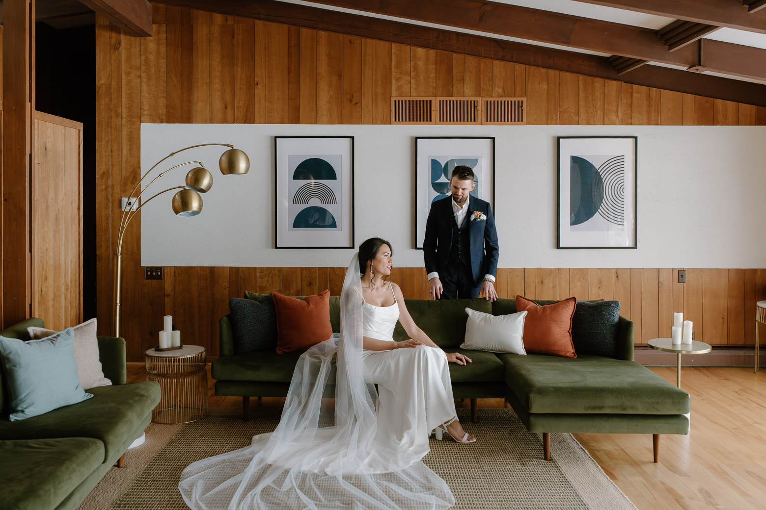 An bride sits on a velvet couch inside a mid-century modern designed living area, and groom stands behind the couch during an elopement at MidMod Minnesota Lakehouse in Grand Rapids, MN.